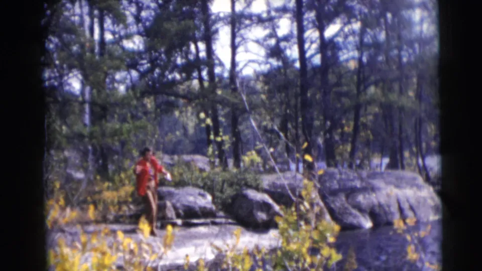1953 Baraboo, Wisconsin home-movie frame showing a man in a red jacket fishing by lakeside rocks in an autumn forest.