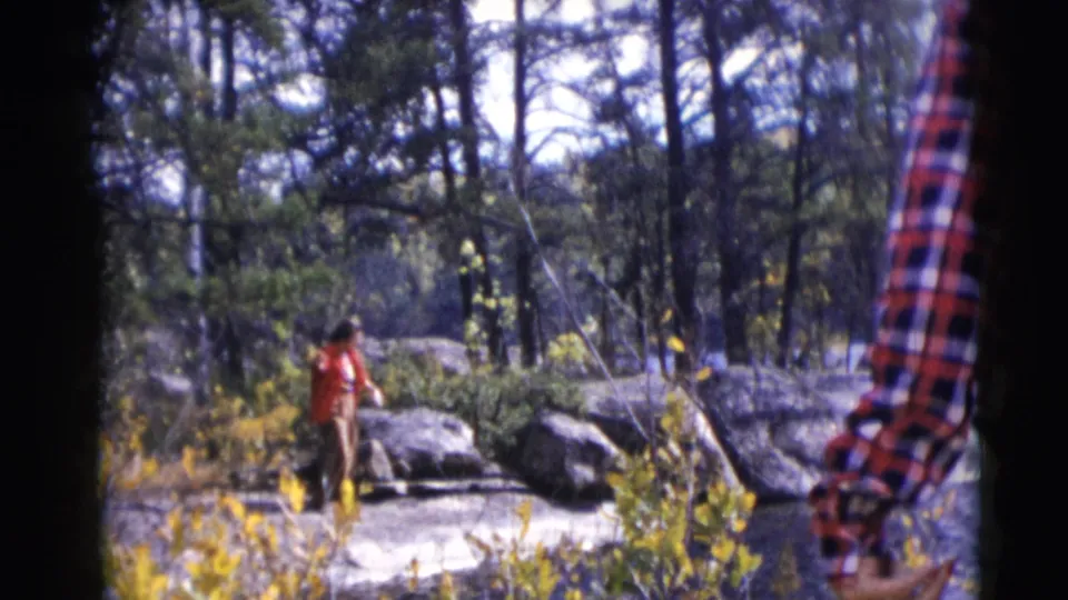 A man fishes by a rocky lakeshore in Baraboo, Wisconsin, while a plaid-shirted person is partly visible in the foreground.