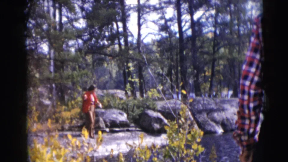 Home-movie view of a man fishing on a wooded Wisconsin lakeshore, with autumn brush and a plaid-shirted person partly blocking the right edge.