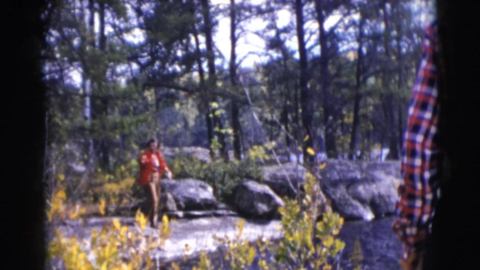 1953 Wisconsin 8mm frame of a man in a red jacket fishing near large rocks, lake water, pine trees, and autumn foliage.