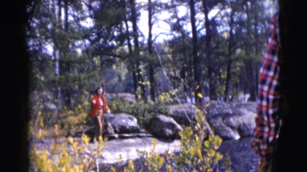 Woman in a red coat stands near grey rocks in a wooded area.