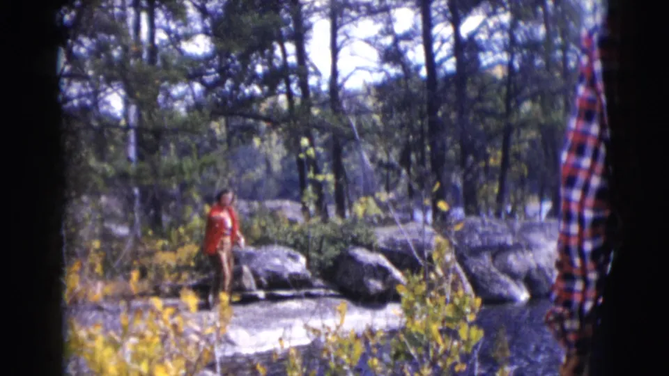 The woman in the red coat remains near large grey rocks in wooded Baraboo, with a plaid sleeve close to the camera edge.