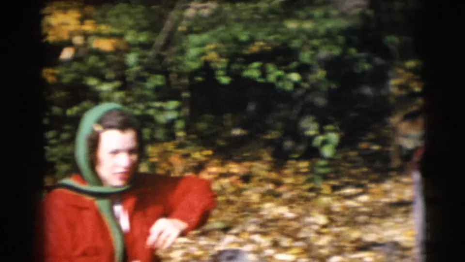 A closer 1953 home-movie frame shows the woman in a red coat and green headscarf seated or crouched near autumn leaves.