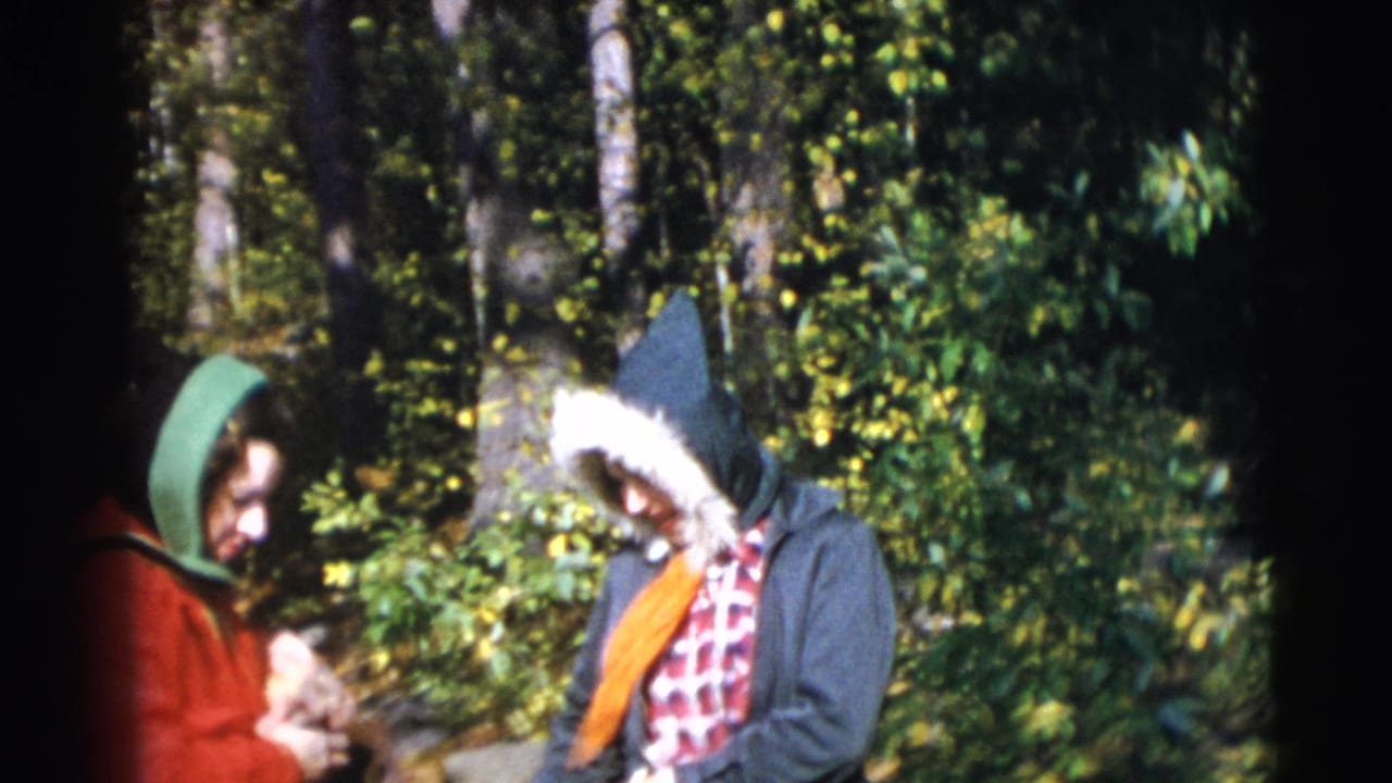 Two hooded women stand together in dense woods.