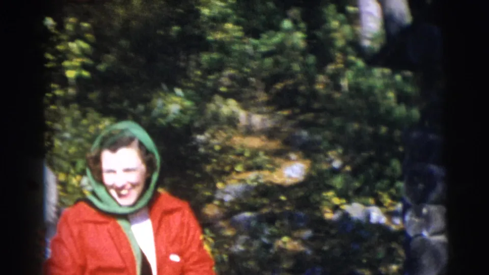 A woman in a red coat and green hood smiles toward the camera in 1953 Baraboo home-movie footage.