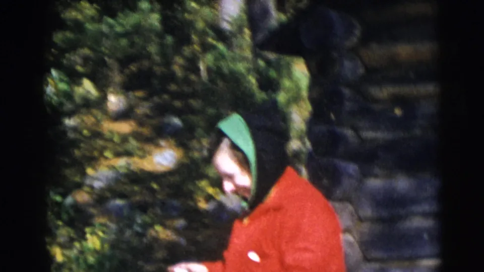 A hooded woman in a red coat stands beside a rustic log structure in the wooded Baraboo setting.
