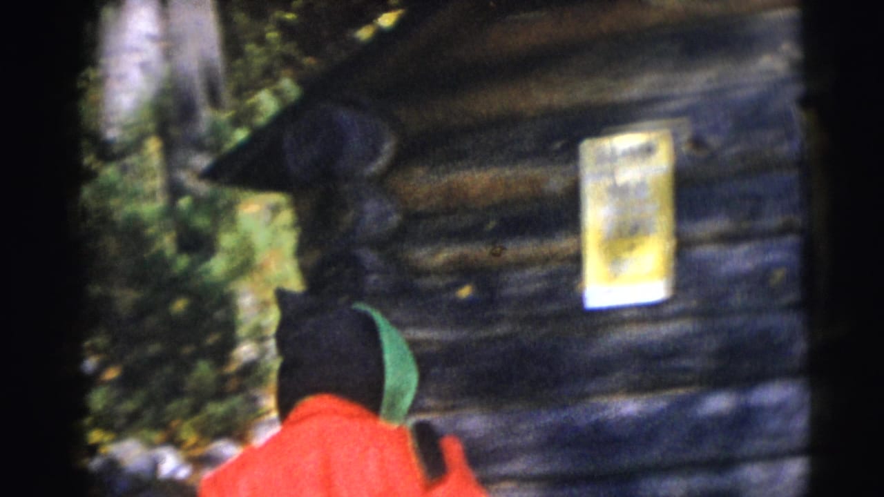 1953 Baraboo home-movie frame showing a person in a red jacket and green hood beside a rustic log cabin wall.