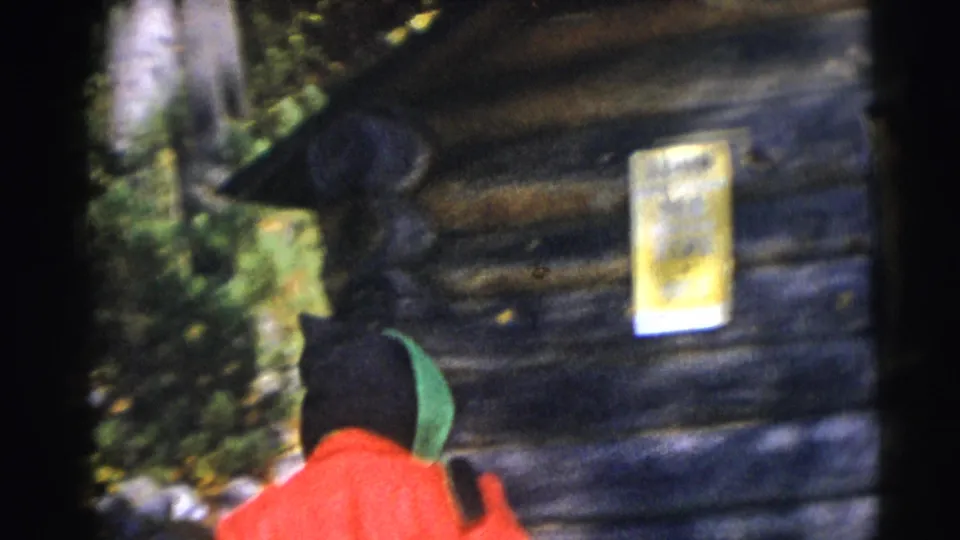 1953 Baraboo home-movie frame showing a person in a red jacket and green hood beside a rustic log cabin wall.