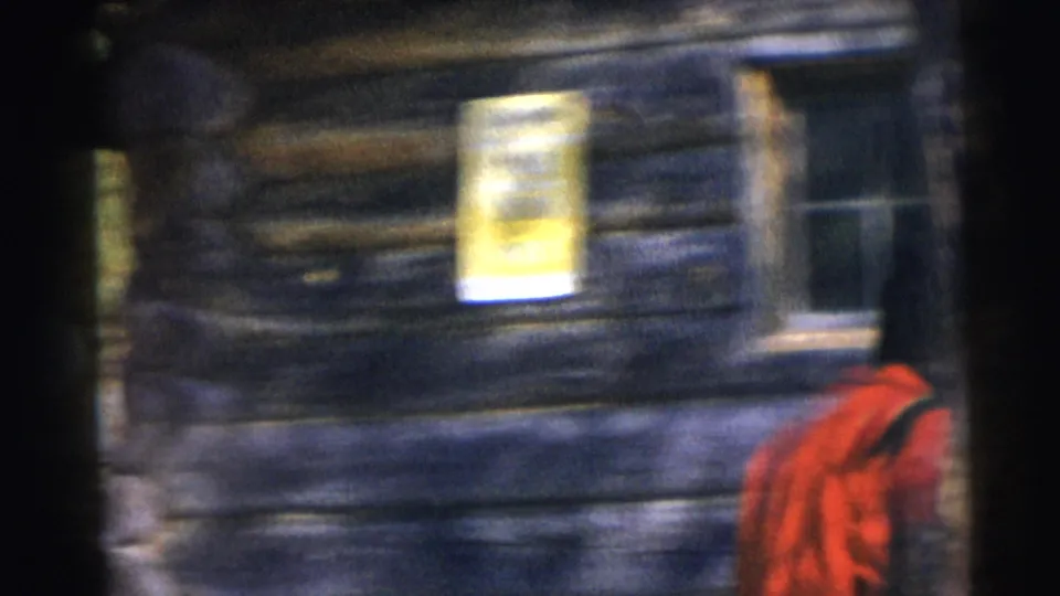 Soft 8mm frame of the Baraboo log cabin exterior, with a yellow posted sign and a red-coated figure partly visible.