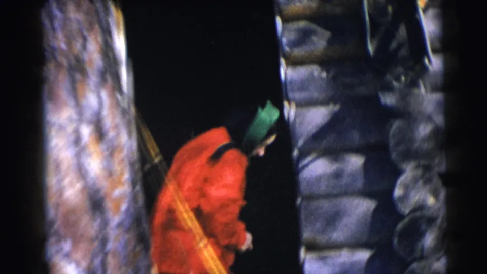 A person in a red jacket bends toward the dark entrance of a rustic log cabin in the 1953 Wisconsin footage.