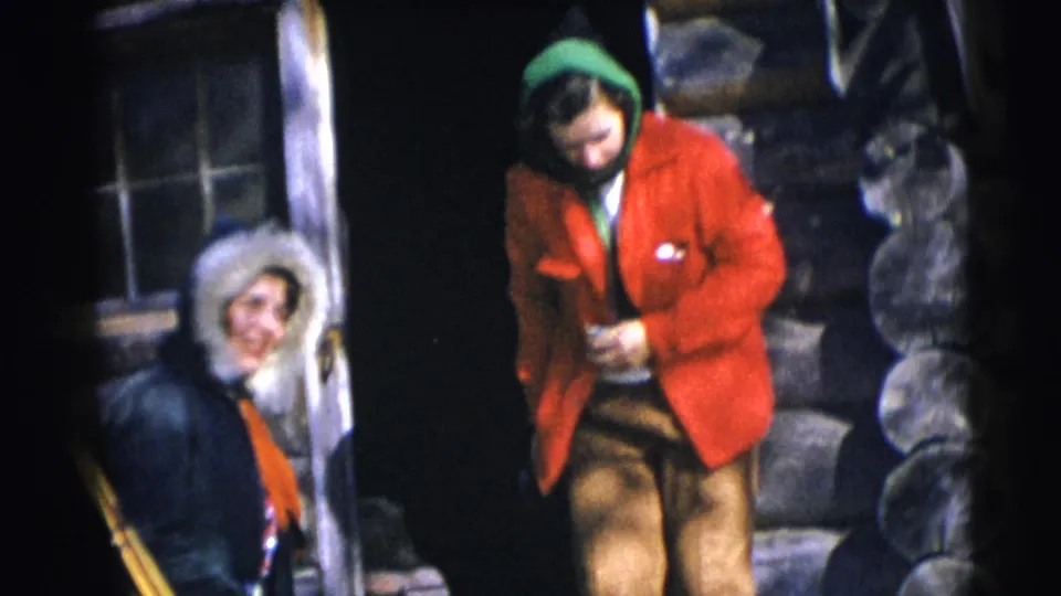 1953 Baraboo frame showing two people at a rustic cabin entrance, including a smiling figure in a fur-trimmed hood.
