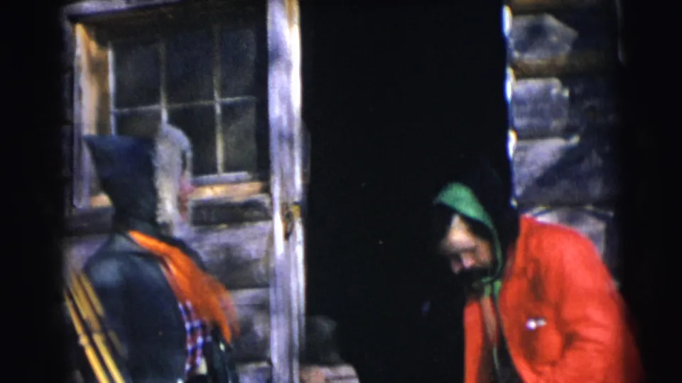 In Baraboo, Wisconsin, 1953 home-movie footage, two adults stand near a log cabin doorway, one in a fur-trimmed hood and orange scarf.