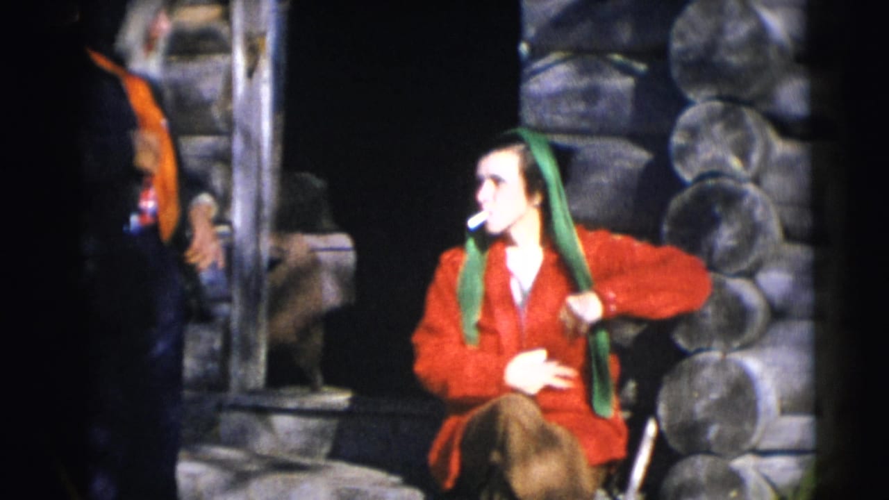 Woman in a red jacket and green headscarf sitting beside a log cabin doorway.