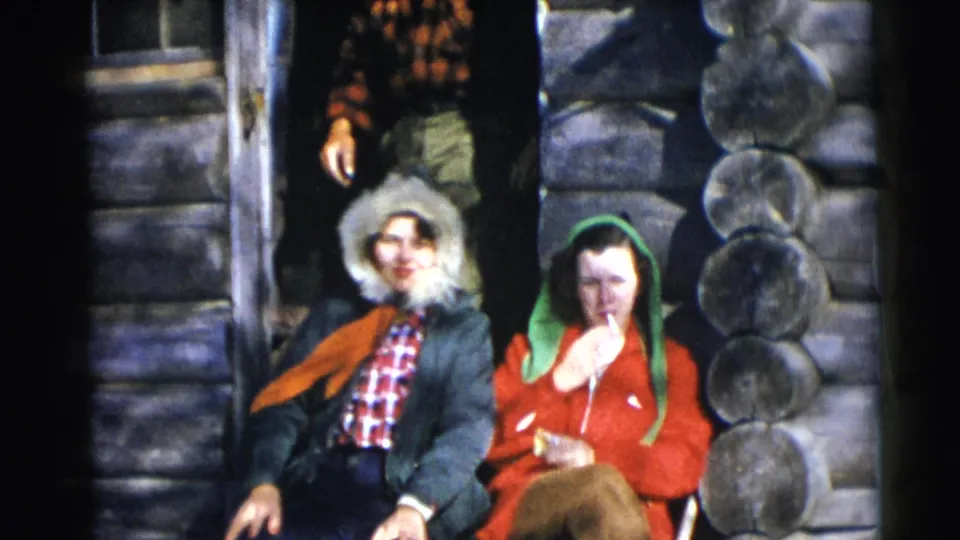 Two women sit at a log cabin doorway in Baraboo, Wisconsin, with winter clothing and casual home-movie gestures visible.