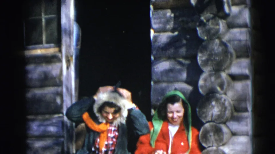 Two women outside a Baraboo log cabin smile toward the camera as one adjusts a fur-trimmed hood in 1953 8mm footage.