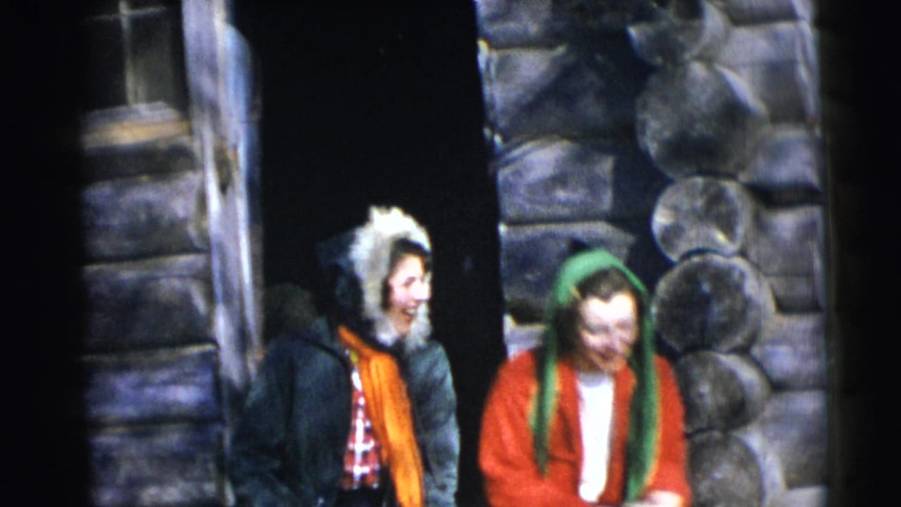 Two young women in winter coats sit by a log cabin doorway and laugh together.