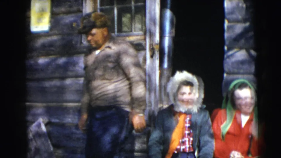 A man in a cap and work clothes passes in front of two seated young women outside the rustic cabin during the 8mm home-movie scene.