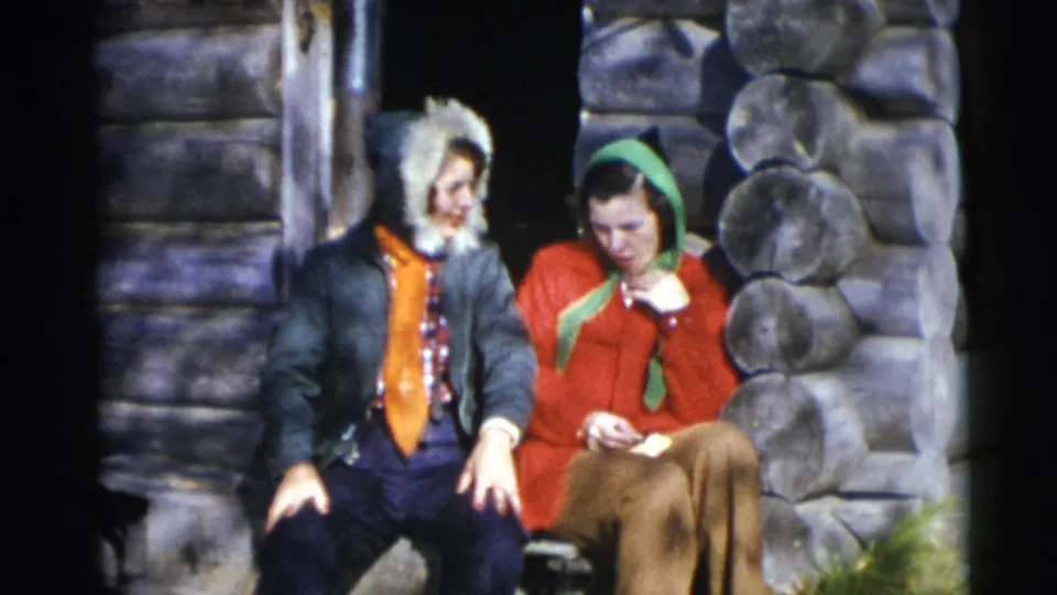 Two young women sit closely on the cabin steps in supported 1953 Baraboo footage, with one looking down at a small item in her hands.