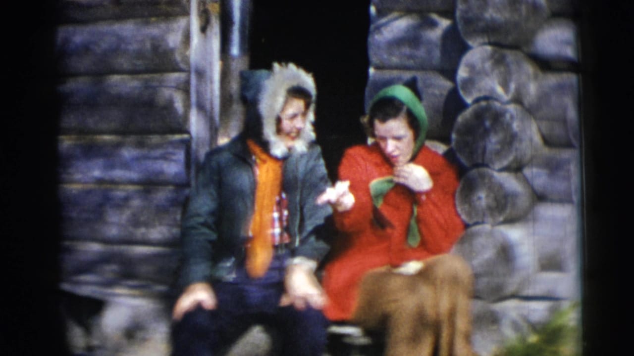 Two adults sit outside a log cabin, with a brown animal partly visible in front.