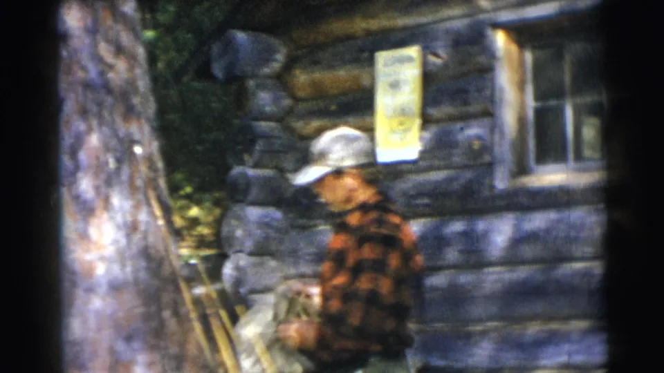 A man in a plaid shirt walks past the log cabin exterior, with trees, a window, and a posted sign visible nearby.