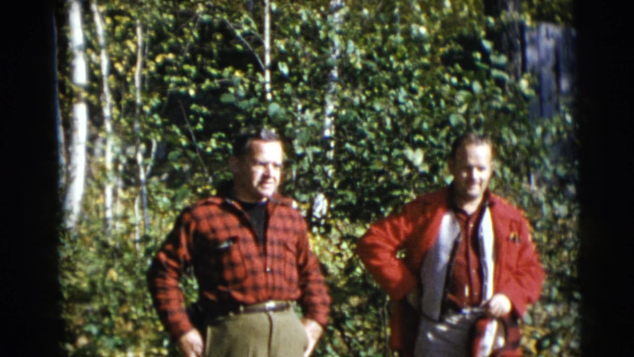 Two adult men in red clothing stand in front of green trees and birch trunks.