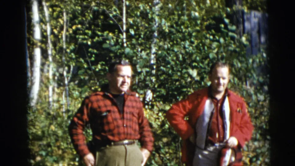 Two adult men stand in sun-dappled woods in Baraboo, Wisconsin, in 1953, one in red plaid and one in a red jacket with a light vest.