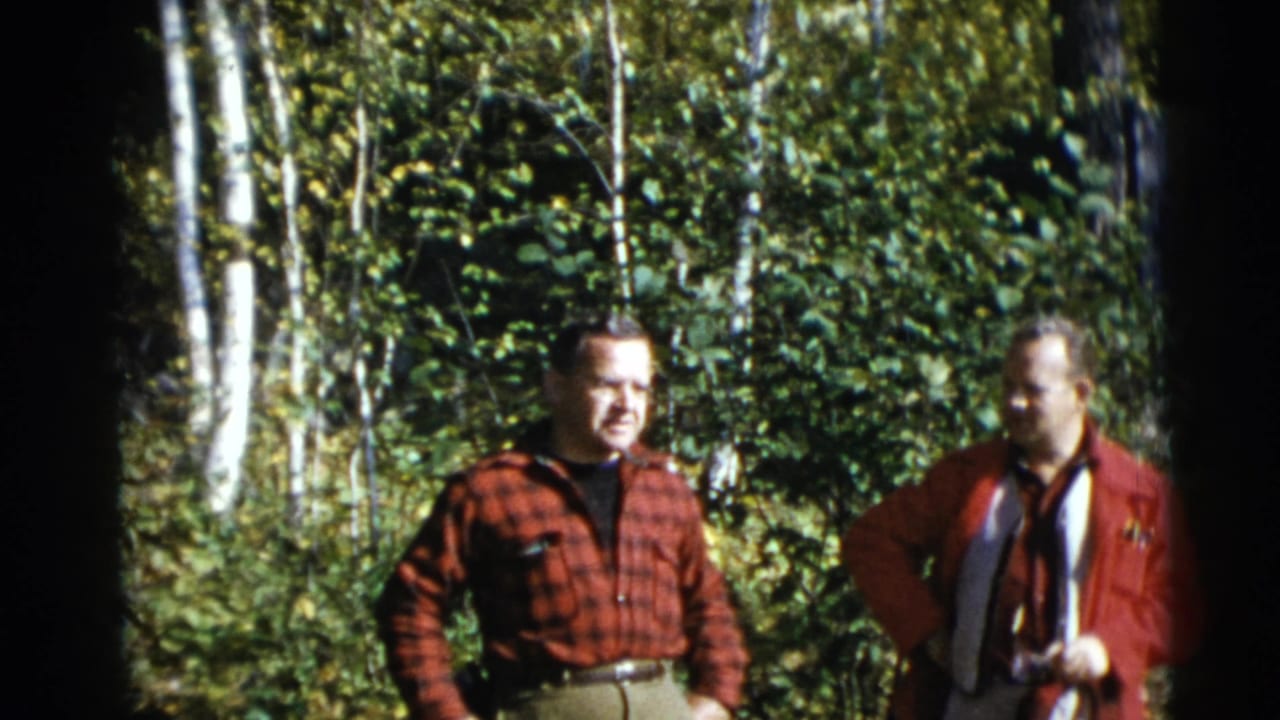 In 1953 Baraboo, Wisconsin footage, two adult men stand near dense green foliage, with white birch trunks visible behind them.