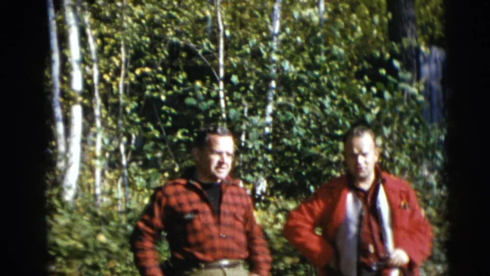 Two adult men in red outdoor clothing stand in a wooded Baraboo, Wisconsin setting, with soft 8mm color and leafy background detail.