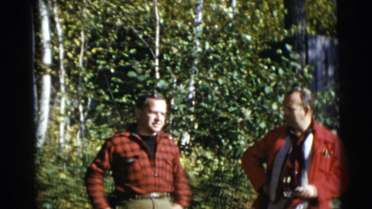 Two adult men in red clothing stand in a sunlit wooded area.