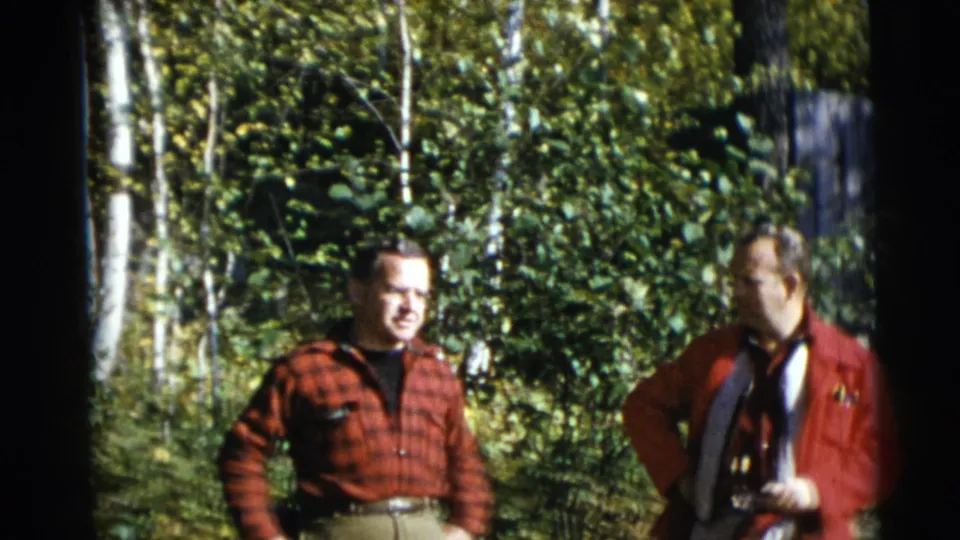 Two adult men stand among green foliage and birch trees in Baraboo, Wisconsin, in 1953 home-movie footage.