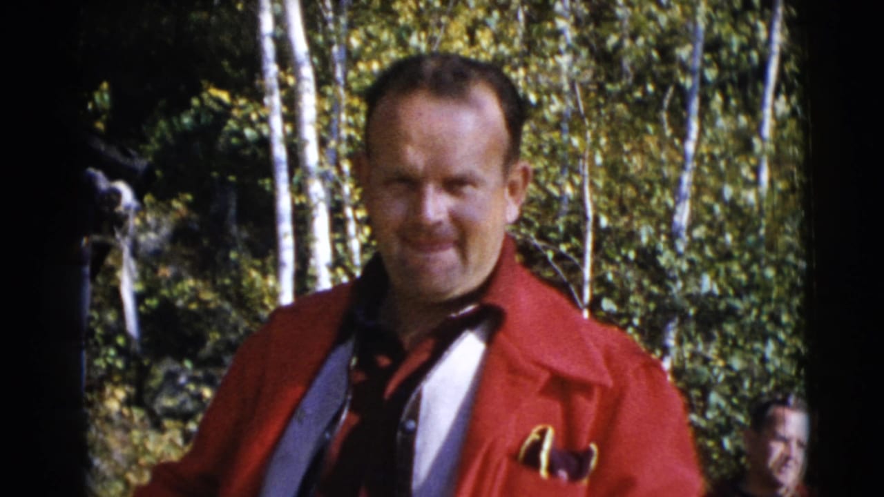 Man in a red jacket standing in sunlit woods with another person partly visible behind him.