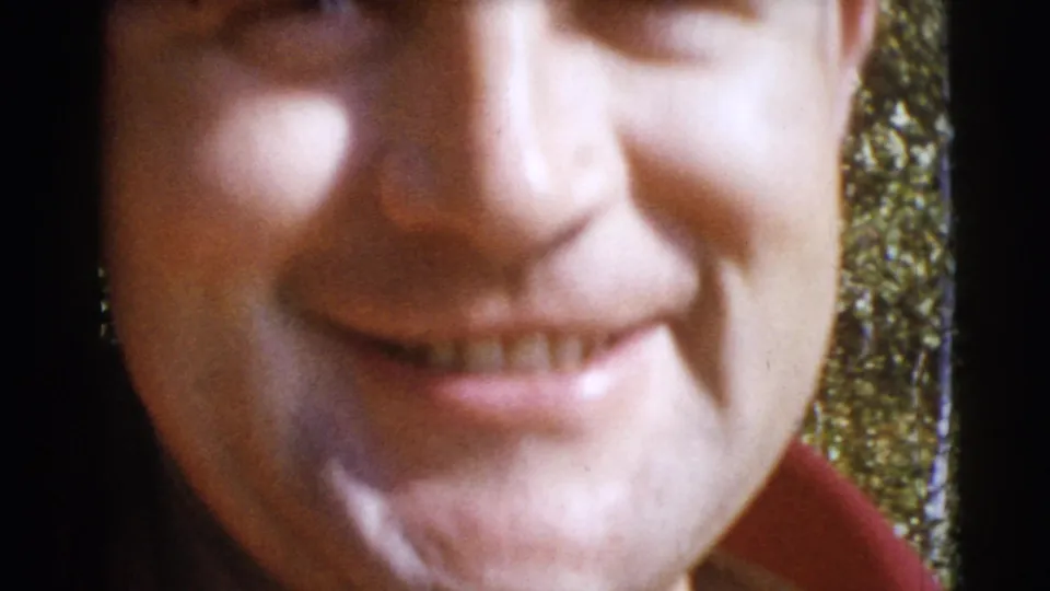 Very close, joking home-movie framing of an adult man's smile, with wooded background visible at the edge.