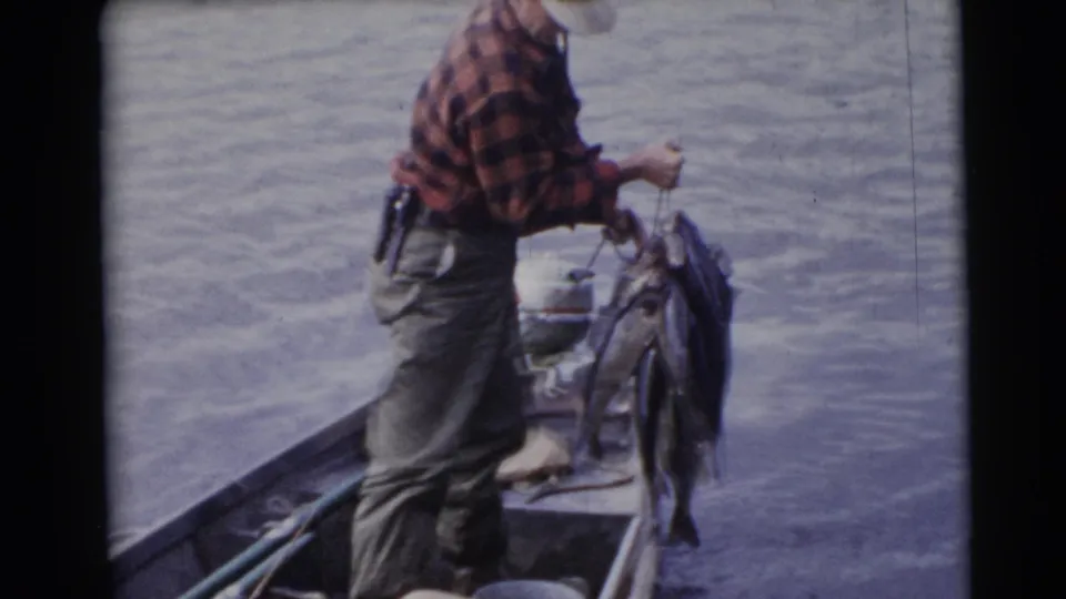 A mid-century home-movie frame shows the fisherman steadying a large group of fish on a stringer inside the small boat.