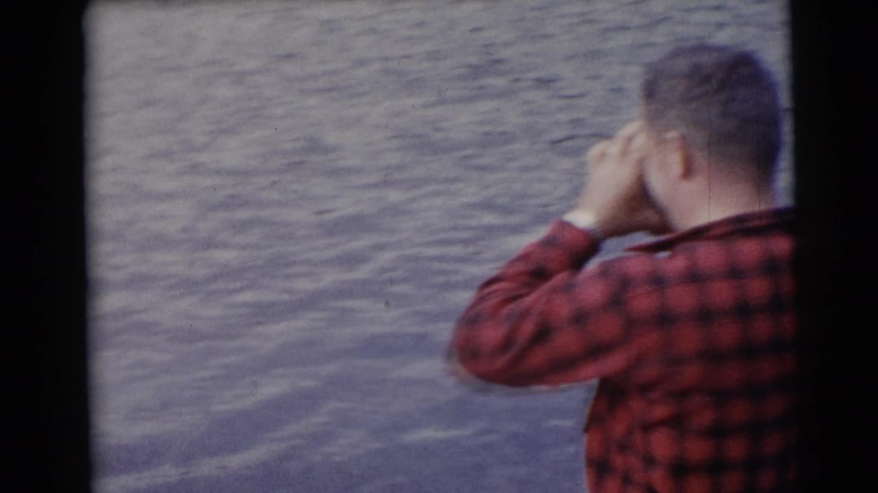 Man in a red plaid shirt stands on a boat facing rippling water.