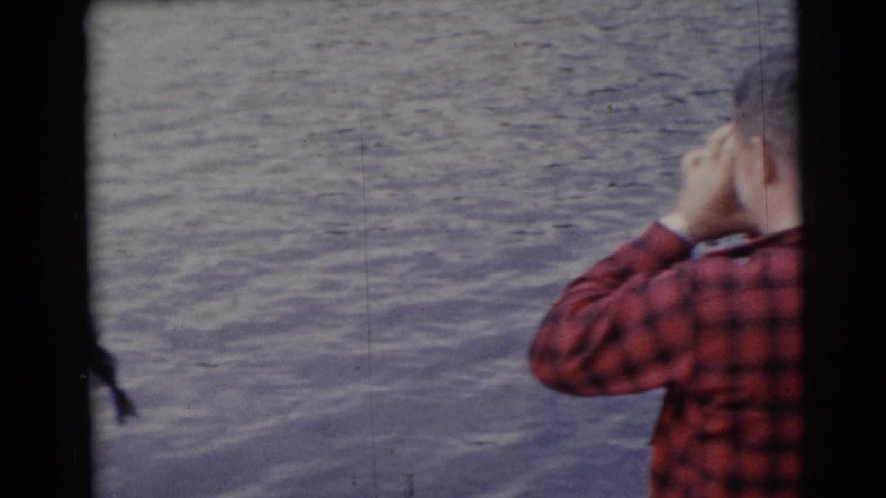 The man in red plaid appears to shield his eyes or adjust something near his face while standing by the water.