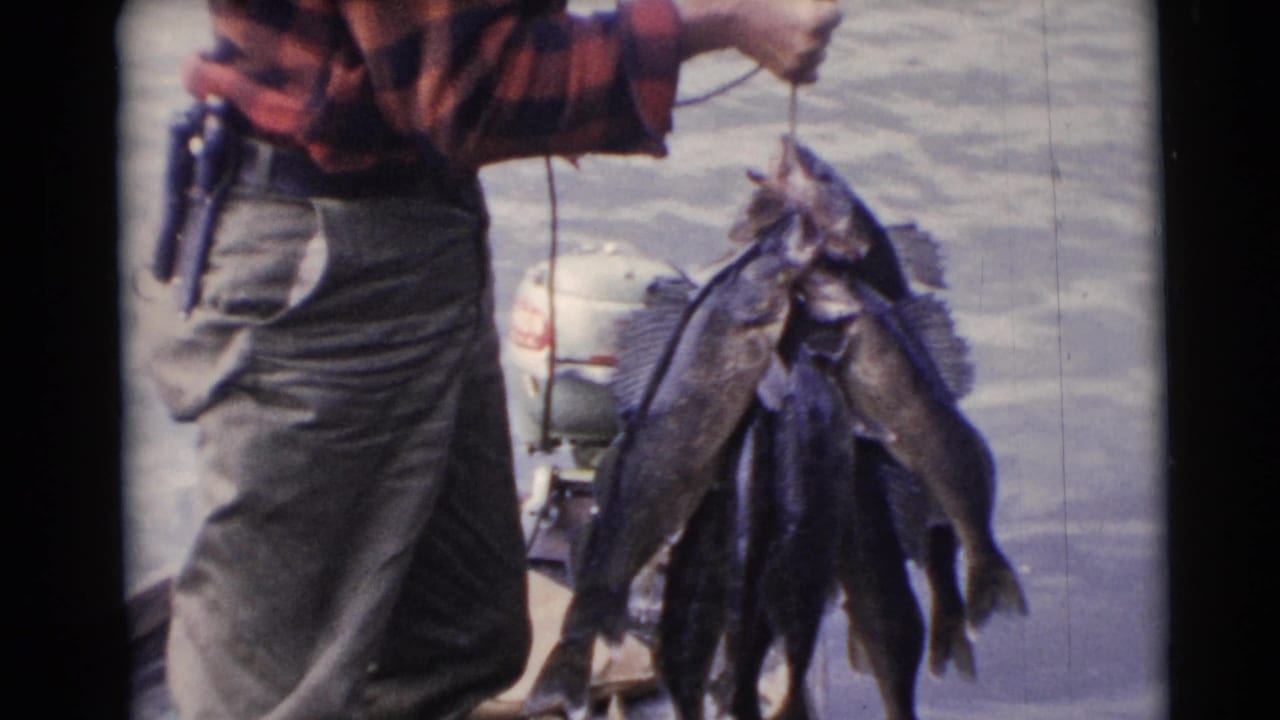 Man in a red plaid shirt holds up a stringer of fish beside water on a small boat.