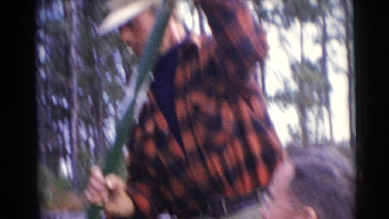 Man in a plaid shirt stands in a small boat holding a green pole near wooded water.