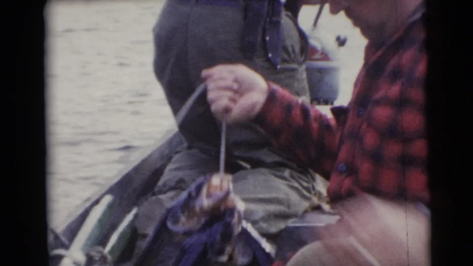 A man in a red plaid shirt sits in the boat holding a rope or line, with water and gear visible in the 1953 Baraboo clip.