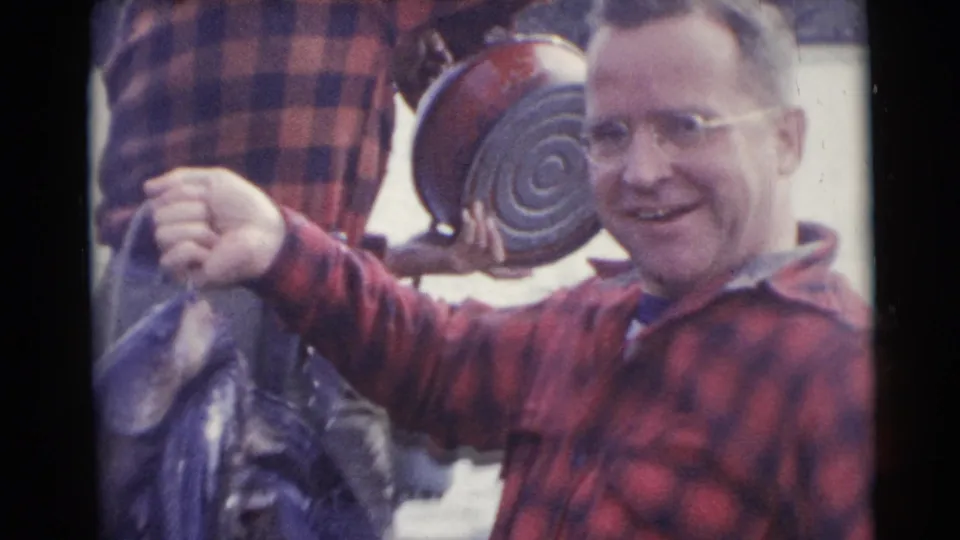 1953 Baraboo, Wisconsin frame of a smiling man in glasses and red plaid holding fish beside a metal pot in a small boat.