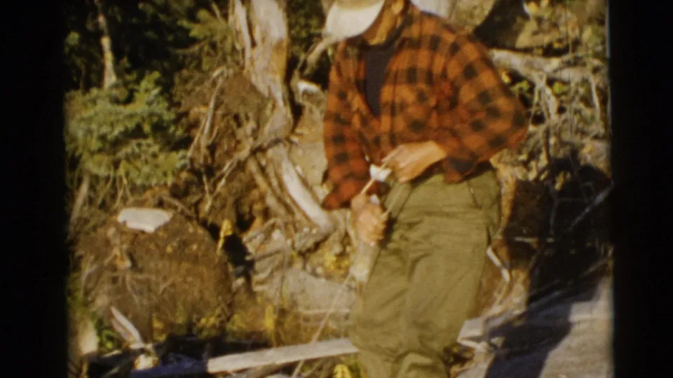 A man in a red plaid shirt, green trousers, and light cap stands near a wooded shoreline in the 1953 Baraboo, Wisconsin fishing footage.