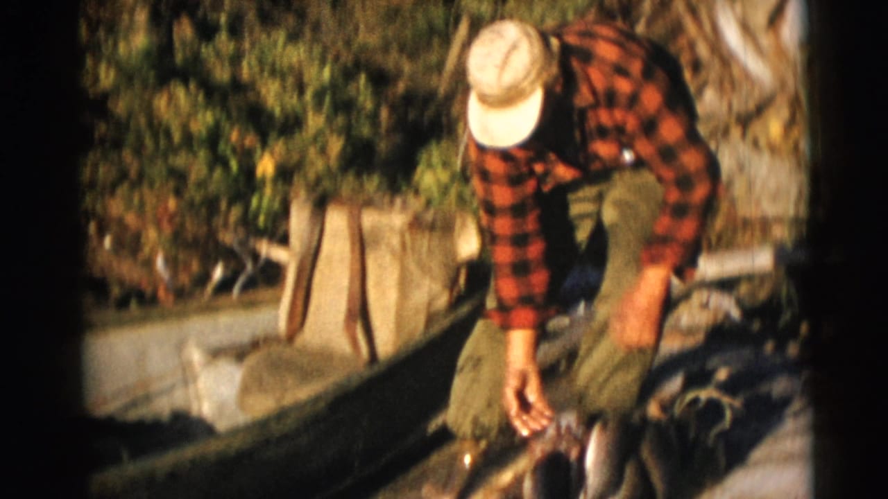 The fisherman reaches down toward several fish beside a wooden boat, with dense greenery behind him.