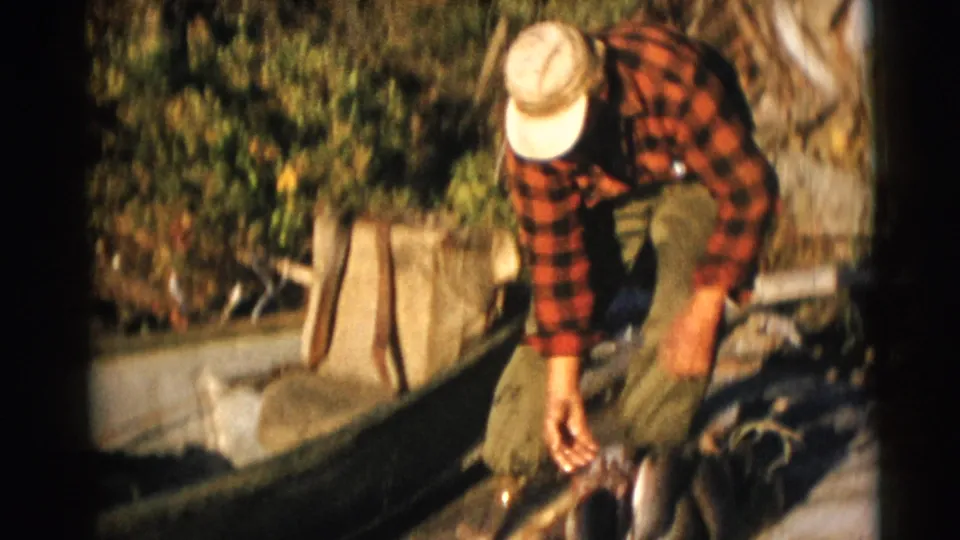 The fisherman reaches down toward several fish beside a wooden boat, with dense greenery behind him.