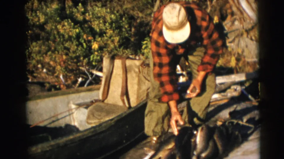 A cap-wearing fisherman stands bent over the catch beside a boat, framed by soft 8mm home-movie exposure.
