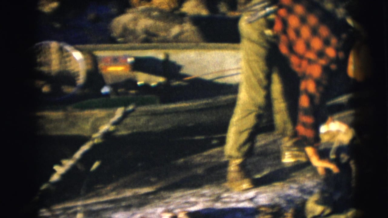 Two men in plaid shirts lean over a small boat near a riverbank.
