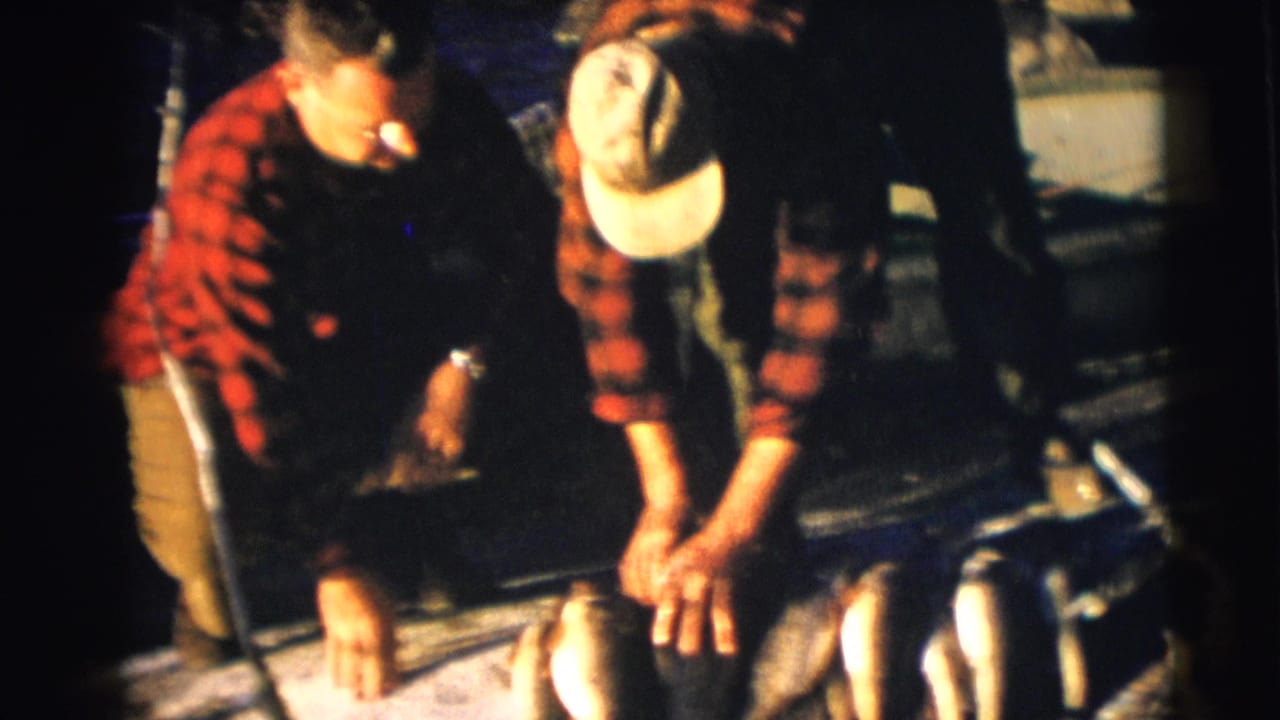 Two men in red plaid shirts crouch beside a boat with a row of fish at the edge of the frame in 1953 Baraboo footage.