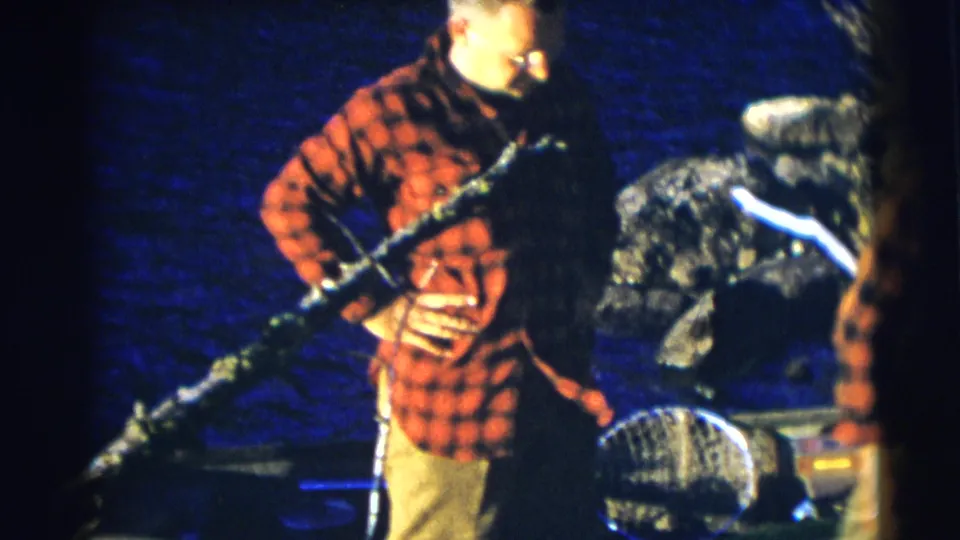 In the Baraboo, Wisconsin fishing scene, an adult man in a red plaid shirt holds a long fishing rod near the boat with water behind him.