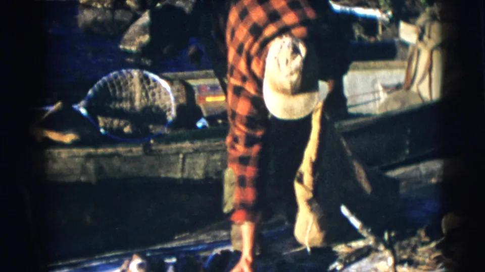 A mid-century 8mm frame shows the Baraboo fisherman bending low in the boat with a landing net behind him and a sack hanging beside him.