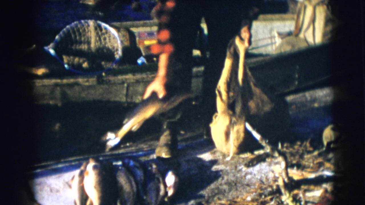 Man in red plaid clothing holds a fish beside a small boat and burlap sack.