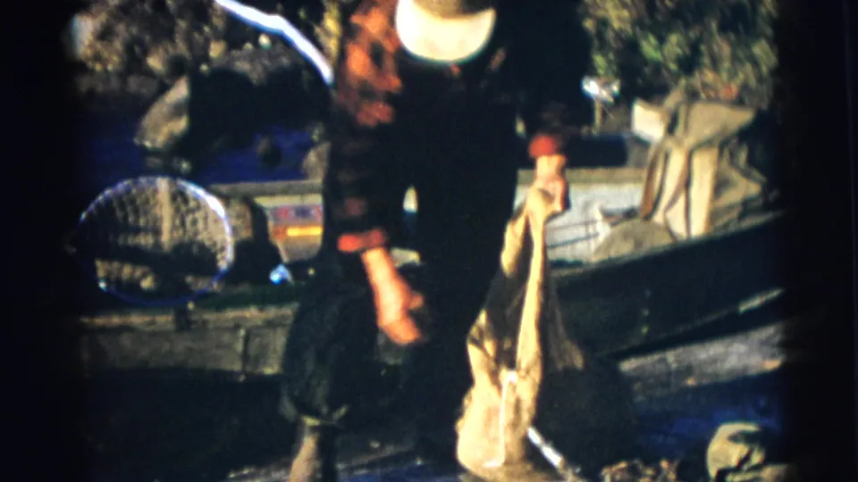 A fisherman in plaid stands at the lakeshore near a small boat, burlap sack, and landing net in 1950s Wisconsin home-movie footage.