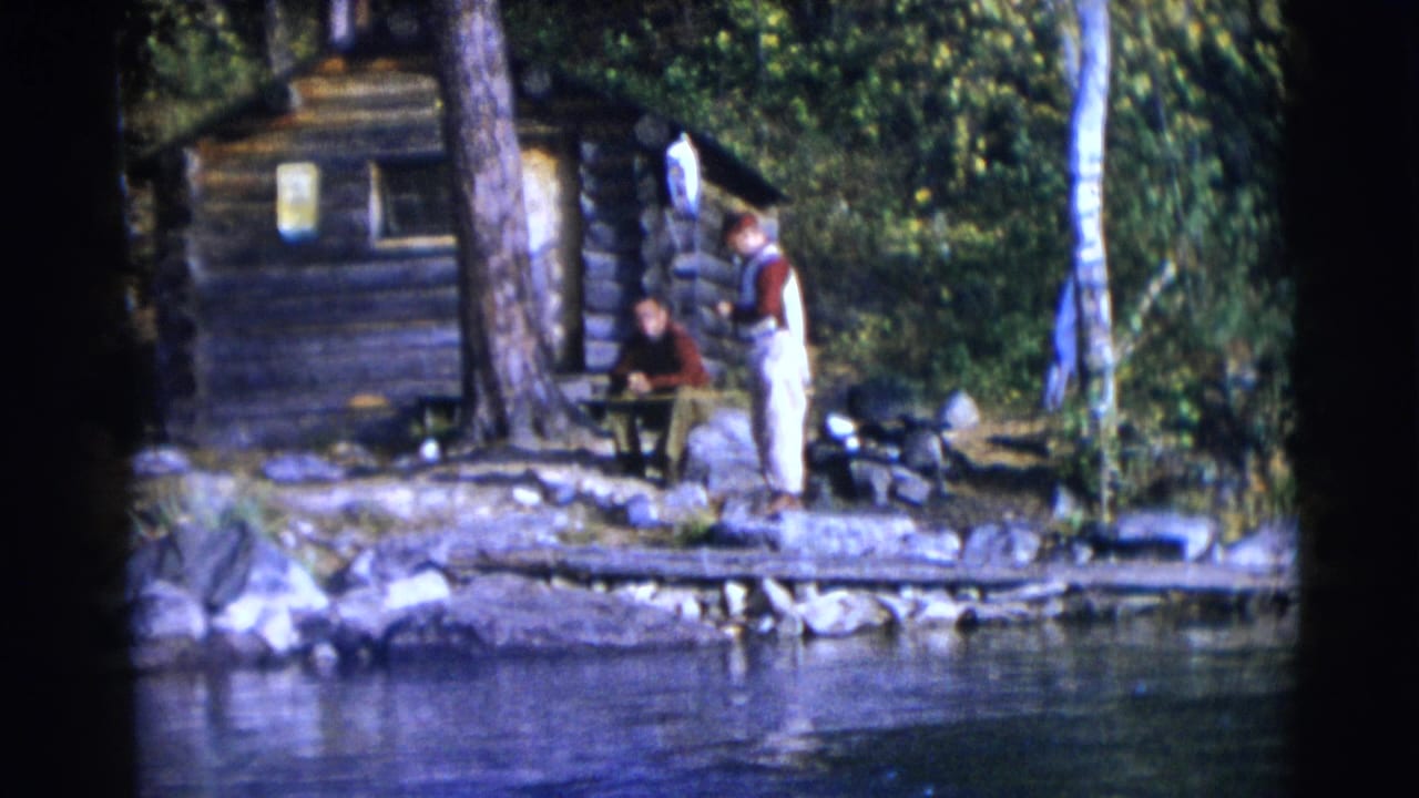 1953 Baraboo home-movie frame showing two men near a lakeside log cabin with trees, rocks, and water in the foreground.